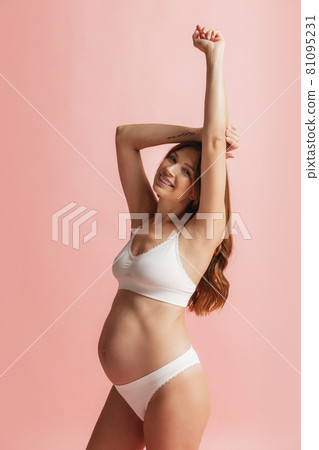 Happy time, awaiting. Portrait of smiling beautiful pregnant woman in lingerie isolated over pink studio background. Natural beauty, happy motherhood, femininity concept. 81095231