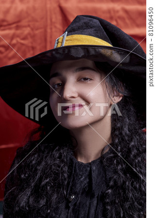 Portrait of a cute multi-racial woman in witch cap against orange background. Studio shot. Close-up shot of female in witch costume 81096450