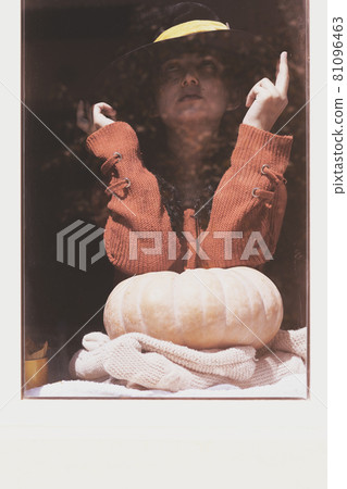 Halloween lady wearing witch cup. A woman in witch hat and Halloween pumpkin near the windows. Portrait of young female in orange sweater and traditional black witch cap 81096463