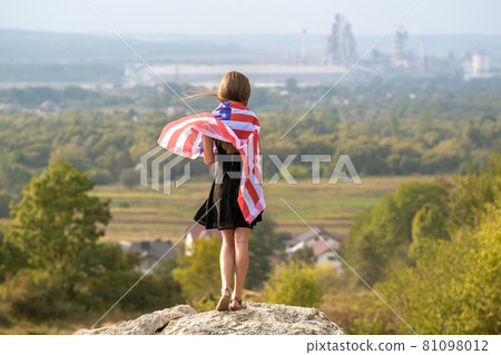 Young happy american woman with long hair holding waving on wind USA national flag on her sholders relaxing outdoors enjoying warm summer day. 81098012