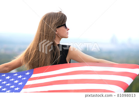 Young pretty american woman with long hair holding waving on wind USA flag on her sholders standing outdoors enjoying warm summer day. Young pretty american woman with long hair holding waving on wind USA flag on her sholders standing outdoors enjoying warm summer day. 81098013