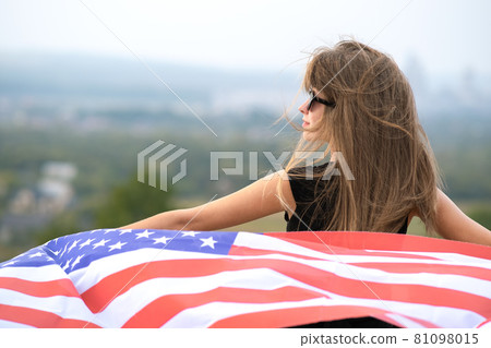Young pretty american woman with long hair holding waving on wind USA  flag on her sholders standing outdoors enjoying warm summer day. 81098015