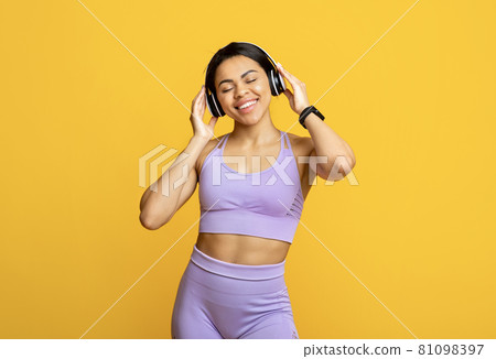 Favourite song. Excited black sportive lady in wireless headphones listening to music and smiling, yellow background 81098397