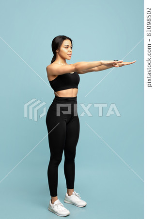 Full length portrait of fit black lady in sports clothes doing squats, exercising on blue studio background Full length portrait of fit black lady in sports clothes doing squats, exercising on blue studio background 81098502