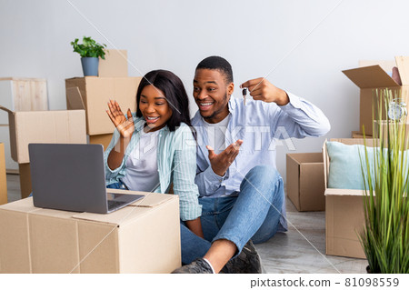 Excited black couple chatting online with friends and showing key of their house to camera, sitting among carton boxes Excited black couple chatting online with friends and showing key of their house to camera, sitting among carton boxes 81098559