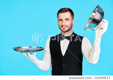 Elegant young waiter holding cloche over empty tray ready to serve on blue background 81099817