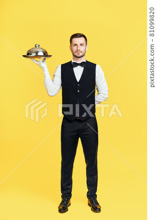Full length portrait of elegant handsome waiter holding tray with lid ready to serve on yellow background 81099820