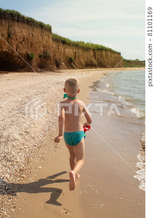 A boy runs along the beach in swimming trunks. He has a water pistol in his hands. Happy child on the sandy beach. Concept of happy life and relaxation by the ocean. Blond boy smiling at the camera A boy runs along the beach in swimming trunks. He has a water pistol in his hands. Happy child on the sandy beach. Concept of happy life and relaxation by the ocean. Blond boy smiling at the camera 81101169