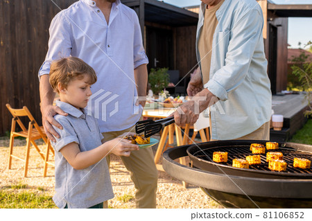 Adorable boy holding plate while his grandfather putting him grilled corn 81106852