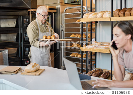 Aged baker with tray putting pastry on display with busy female colleague in front Aged baker with tray putting pastry on display with busy female colleague in front 81106988