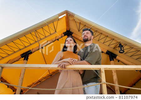 Young serene couple standing by glamping house and looking forwards Young serene couple standing by glamping house and looking forwards 81107974