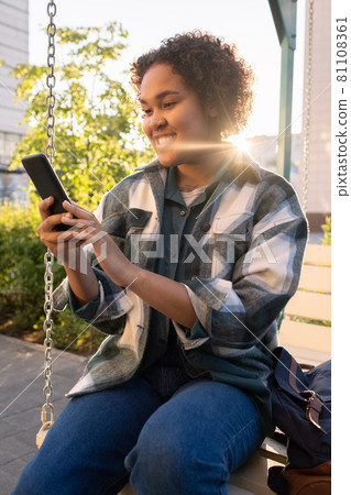 Happy African female teenager with smartphone sitting on swing Happy African female teenager with smartphone sitting on swing 81108361