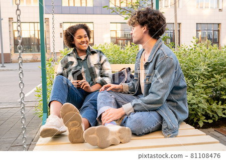 Two carefree teens in casualwear sitting on swings and interacting Two carefree teens in casualwear sitting on swings and interacting 81108476