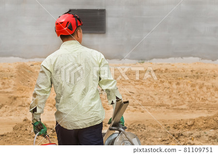 Industrial worker in a red helmet walks with a working tool against the background of a sand and concrete building house at a construction site Industrial worker in a red helmet walks with a working tool against the background of a sand and concrete building house at a construction site 81109751