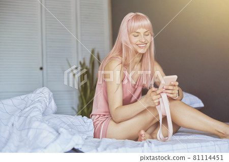 Happy lady with long pink hair holds smartphone sitting on comfortable bed Happy lady with long pink hair holds smartphone sitting on comfortable bed 81114451