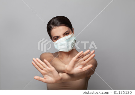 Serious woman in surgical protective face mask showing stop gesture on gray background 81125120