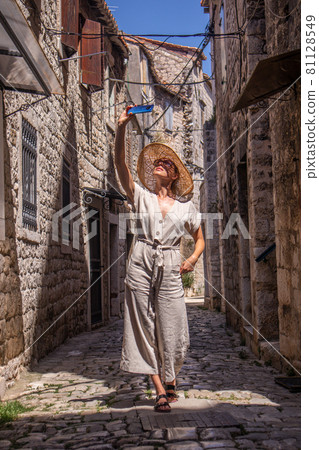 Beautiful blonde young female traveler wearing straw sun hat, taking selfie while sightseeing and enjoying summer vacation in an old traditional costal town at Adriatic cost, Croatia. 81128549