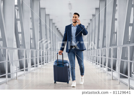 Handsome Arab Man Walking With Suitcase In Airport And Talking On Cellphone 81129641