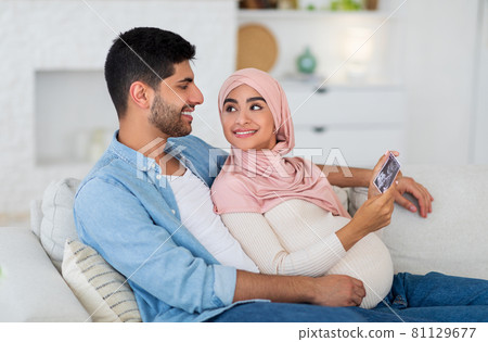 Happy islamic spouses looking at ultrasound photo of their future baby resting at home on sofa, free space Happy islamic spouses looking at ultrasound photo of their future baby resting at home on sofa, free space 81129677