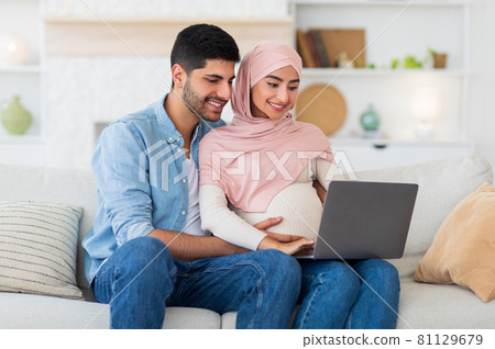 Loving pregnant muslim couple using laptop computer, resting on sofa in living room, spending time together, empty space 81129679