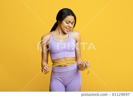 Happy fit young black woman measuring waist with yellow tape and smiling, isolated on yellow background 81129697