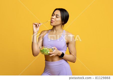 Healthy sporty african american lady enjoying fresh vegetable salad, standing over yellow studio background 81129868