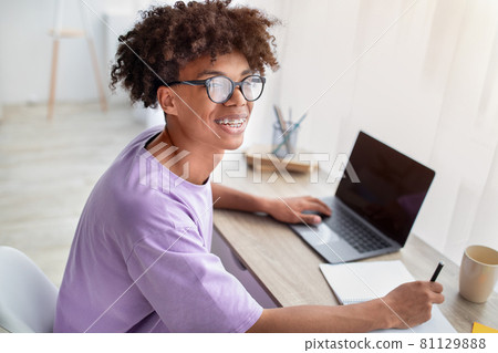 Happy black adolescent sitting at desk with laptop, learning remotely, taking notes in copybook at home 81129888