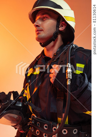 One male firefighter dressed in uniform posing over orange background in neon lights. 81130614