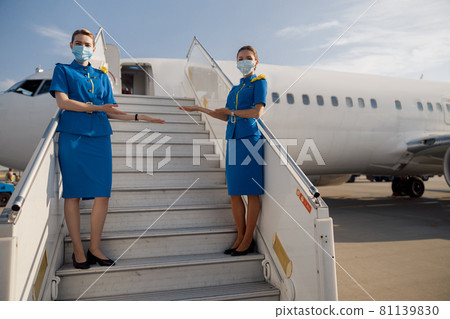 Full length shot of two beautiful air stewardesses in blue uniform and protective face masks looking at camera, standing on airstair and welcoming passengers 81139830