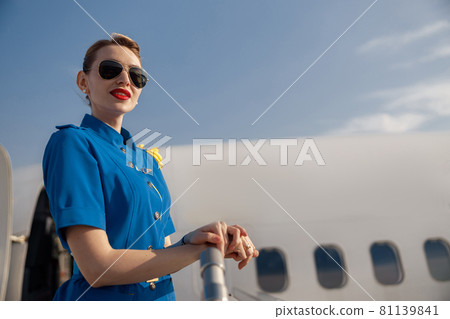 Portrait of pretty air stewardess in blue uniform and sunglasses looking away, standing on airstair on a daytime 81139841