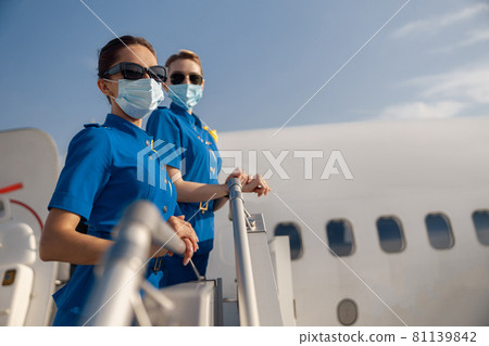 Two young air stewardesses in blue uniform, sunglasses and protective face masks looking at camera, standing on airstair on a daytime 81139842