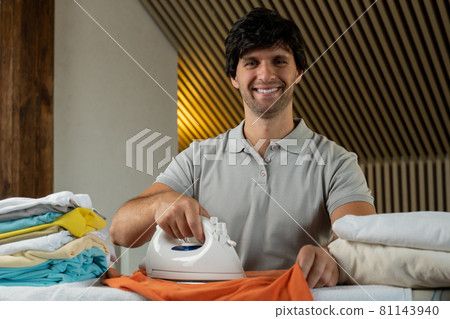 Attractive man is ironing a shirt at home. Portrait of a smiling young man, ironing clothes. 81143940