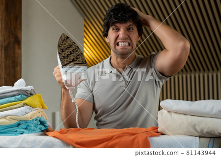 Young man ironing clothes at home angry and mad screaming frustrated and furious, shouting with anger. 81143944