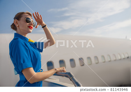 Portrait of elegant air stewardess in blue uniform and sunglasses holding hand near her head and looking far away, standing on airstair on a daytime Portrait of elegant air stewardess in blue uniform and sunglasses holding hand near her head and looking far away, standing on airstair on a daytime 81143946