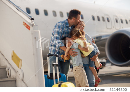 Family of four kissing each other while going on a trip, standing in front of big airplane outdoors Family of four kissing each other while going on a trip, standing in front of big airplane outdoors 81144800