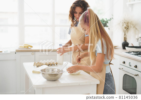 Teen girl helping her mom to cook dough in their kitchen at home Teen girl helping her mom to cook dough in their kitchen at home 81145036