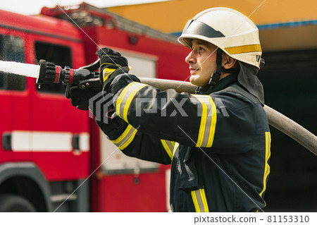 One young male firefighter on duty dressed in uniform with water hose extinguishing fire 81153310