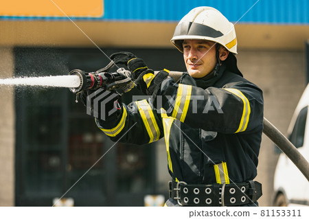 One young male firefighter on duty dressed in uniform with water hose extinguishing fire 81153311