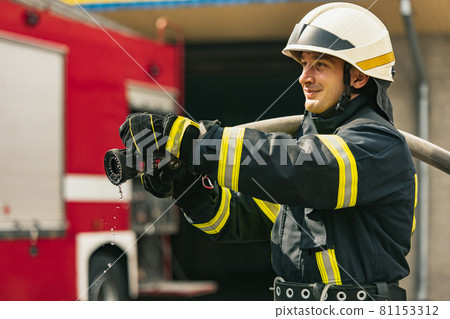 One young male firefighter on duty dressed in uniform with water hose extinguishing fire 81153312