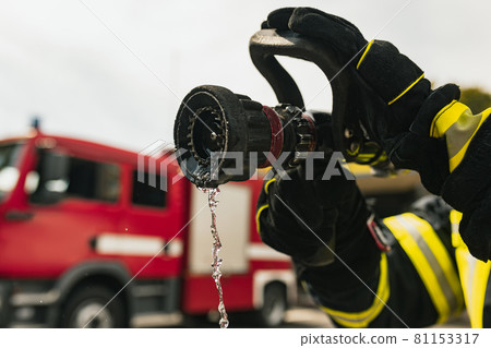 Firefighter on duty with water hose in front of fire truck ready for extinguishing. Close-up Firefighter on duty with water hose in front of fire truck ready for extinguishing. Close-up 81153317