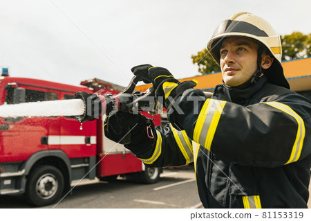 One young male firefighter on duty dressed in uniform with water hose extinguishing fire One young male firefighter on duty dressed in uniform with water hose extinguishing fire 81153319