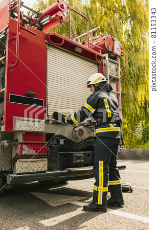 One young male firefighter dressed in uniform with protective helmet near fire engine. 81153343