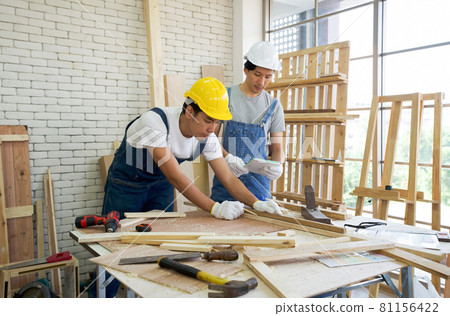 An Asian carpenter is drawing a line on a piece of wood, preparing to cut into sections. The other man told the size he wanted by looking at his note. Morning work atmosphere in the workshop room. An Asian carpenter is drawing a line on a piece of wood, preparing to cut into sections. The other man told the size he wanted by looking at his note. Morning work atmosphere in the workshop room. 81156422