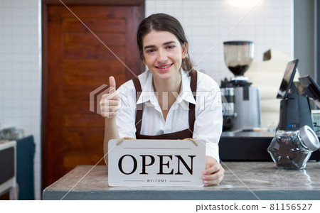Young caucasian shopkeeper with a smile, lift a finger thumb up while holding an OPEN sign in front of a coffee shop counter. 81156527
