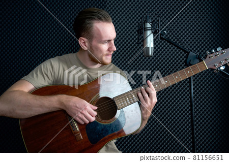 Young caucasian man sings while playing an acoustic guitar in front of black soundproofing walls. Musicians producing music in professional recording studio. Young caucasian man sings while playing an acoustic guitar in front of black soundproofing walls. Musicians producing music in professional recording studio. 81156651