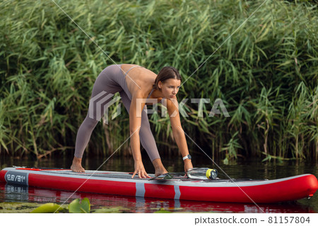 Young Caucasian beautiful slim girl doing yoga, pilates on paddle board, SUP alone in summer morning. Active life, sport, leisure activity concept 81157804