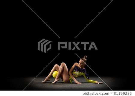 Portrait of little girl, rhythmic gymnastics artist in bright stage costume isolated on dark studio background in ray of light. Concept of sport, action, aspiration, active lifestyle 81159285