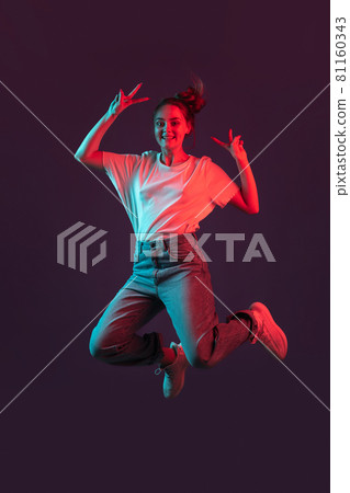 Vertical portrait of beautiful girl in casual clothes jumping isolated on dark purple studio background in red neon light, filter. Concept of human emotions, youth culture Vertical portrait of beautiful girl in casual clothes jumping isolated on dark purple studio background in red neon light, filter. Concept of human emotions, youth culture 81160343