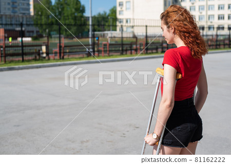 Young red-haired woman in shorts moves with crutches outdoors. 81162222