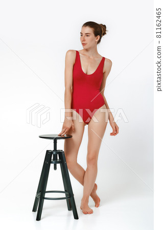 Woman in red body swimming suit posing in studio with a chair Woman in red body swimming suit posing in studio with a chair 81162465